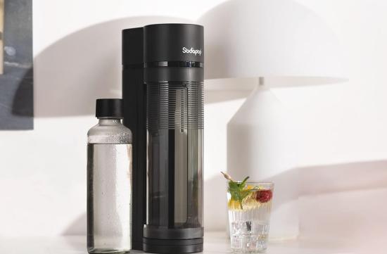 Ein eleganter schwarzer Wassersprudler steht auf einem Tisch neben einer Glasflasche und einem erfrischenden Getränk mit Minze und Beere.