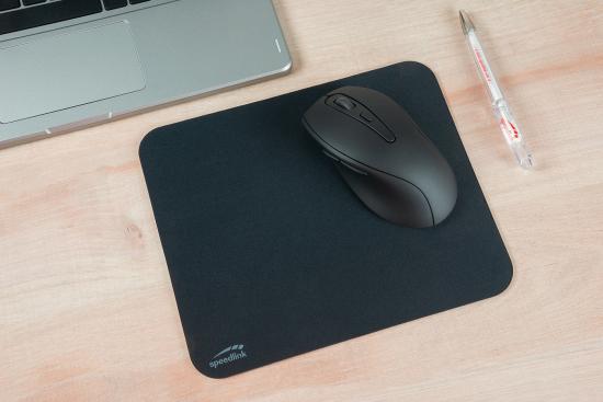 Draufsicht einer ergonomischen schwarzen Maus auf einem Mauspad, platziert auf einem Holztisch neben einem Laptop und einem Kugelschreiber.