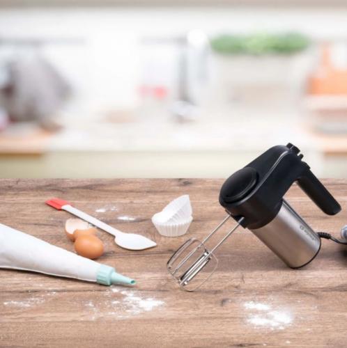 Handmixer auf Holztisch mit Backzubehör wie Spritzbeutel, Eier, Teigschaber und Muffinförmchen in einer Küchenumgebung